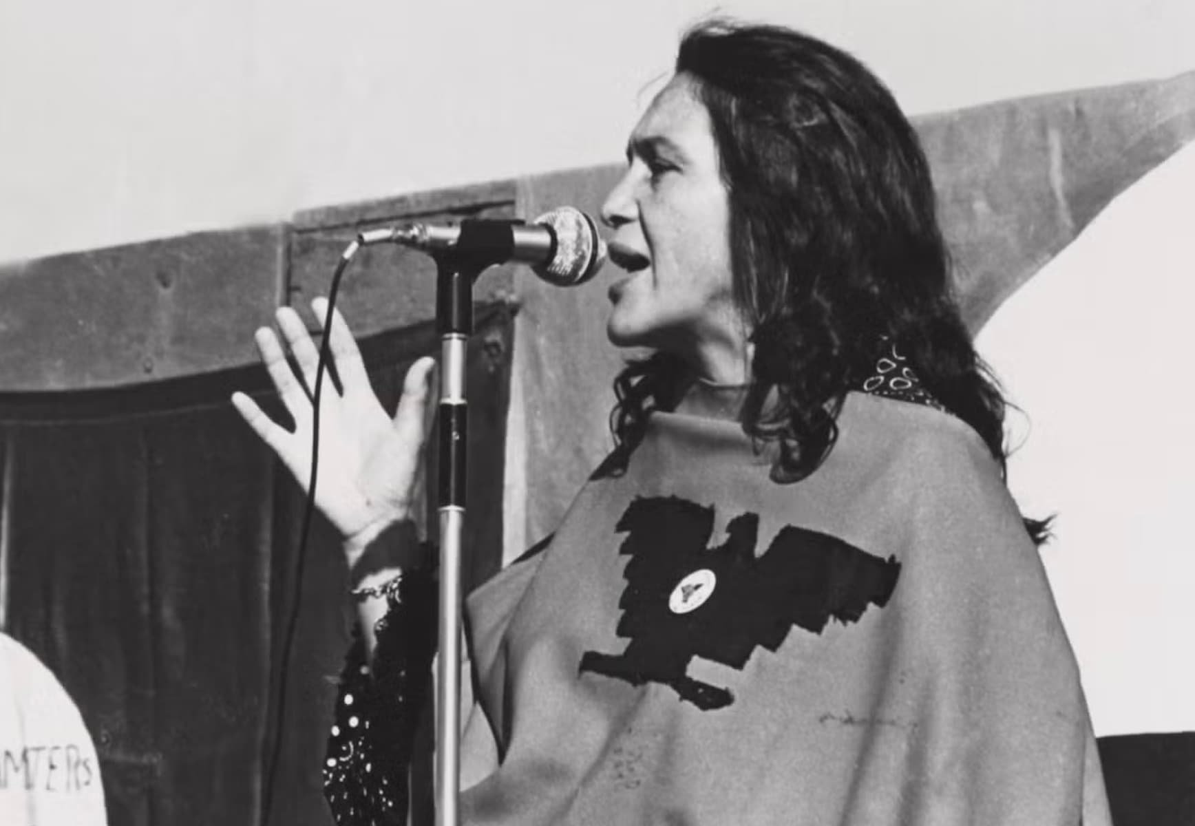 Dolores Huerta speaking into a microphone at a United Farm Workers rally, wearing the UFW eagle poncho