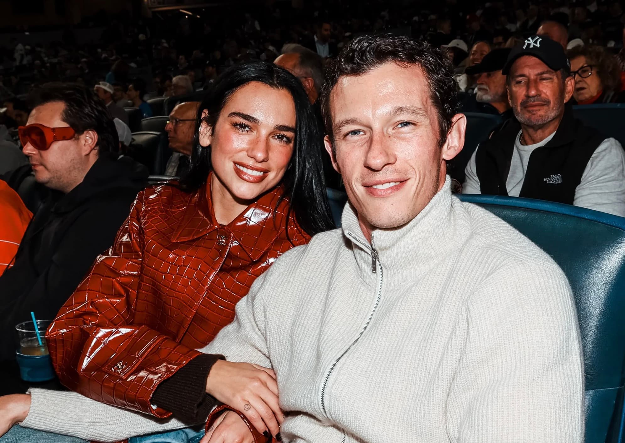 Dua Lipa in an orange croc embossed jacket and Callum Turner in a cream sweater at a baseball game