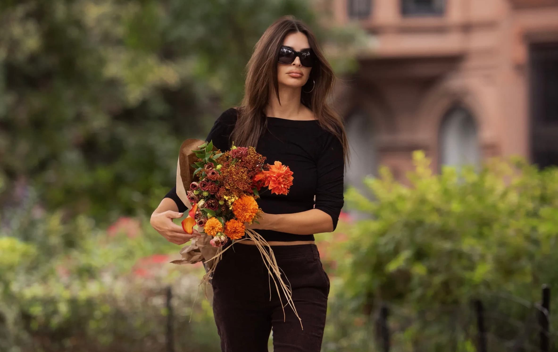Emily Ratajkowski walking outdoors in a black top and brown corduroy pants carrying a bouquet of flowers