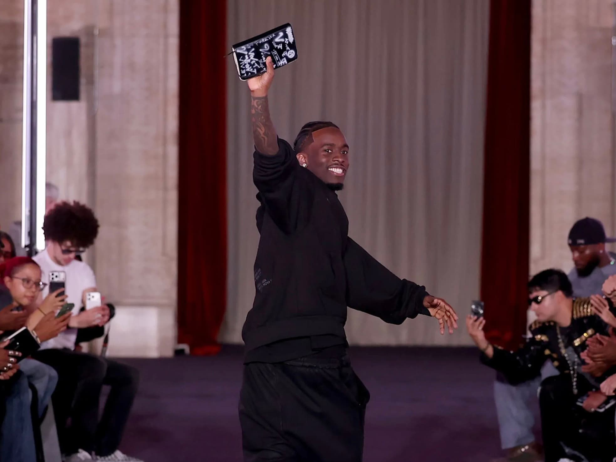 A man celebrating at the end of a runway show, holding up a graffiti covered clutch as the crowd films on their phones