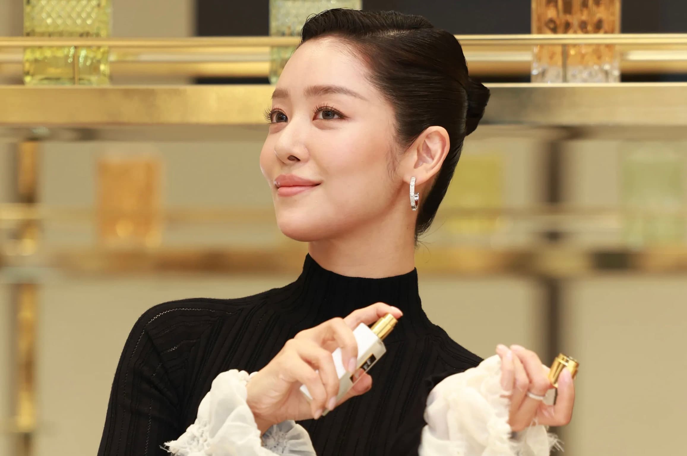 Woman in a black turtleneck holding two Kilian Paris fragrance bottles at a mirrored perfume counter