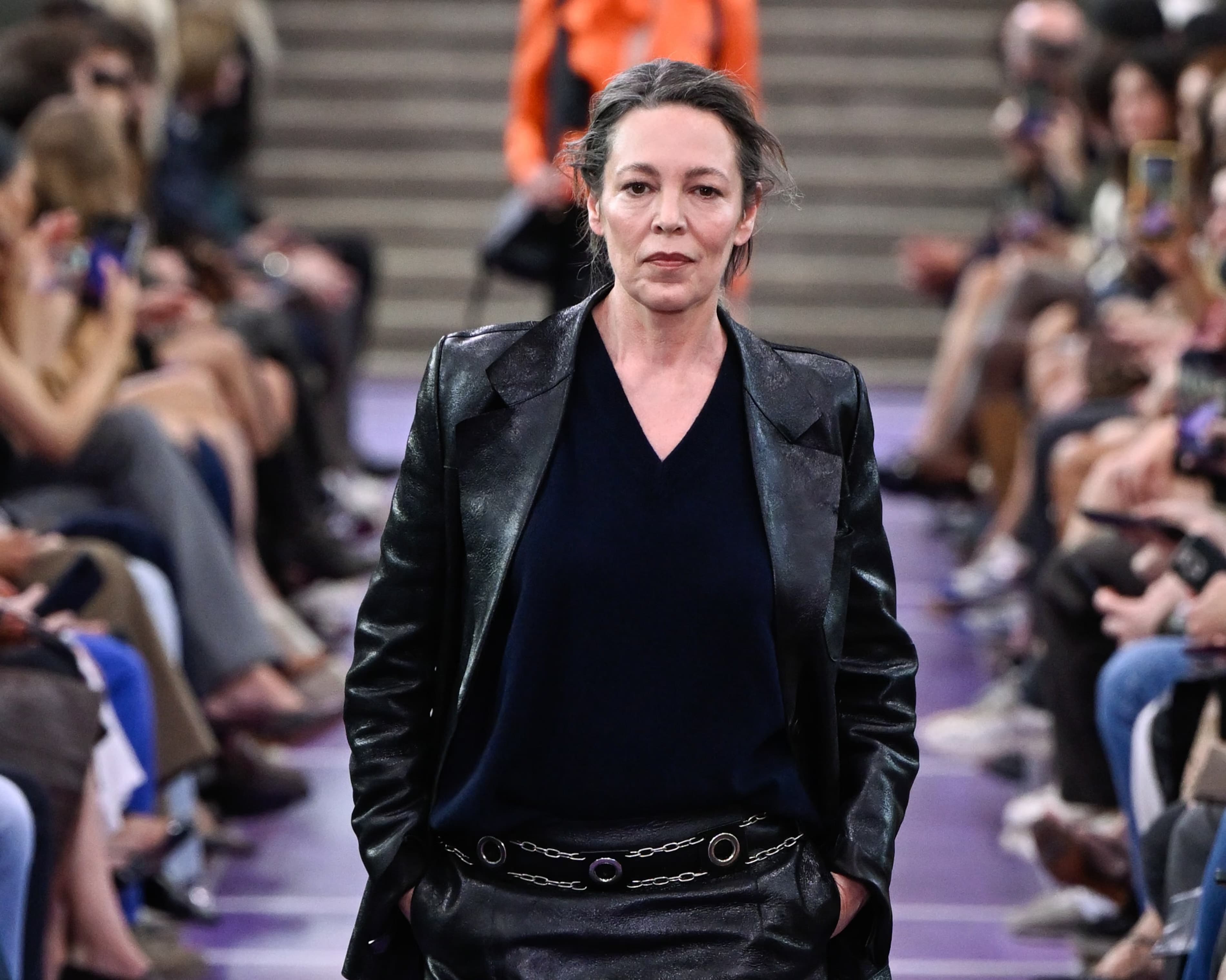 Olivia Colman walking the Miu Miu runway in a black leather blazer, navy sweater, and leather skirt with a chain belt