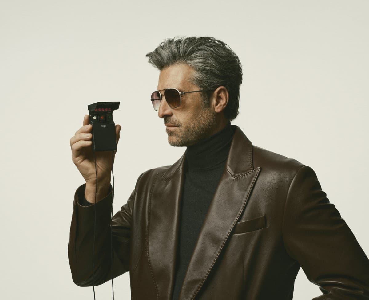 Patrick Dempsey in a brown leather blazer and black turtleneck wearing TAG Heuer sunglasses and holding a vintage Heuer stopwatch