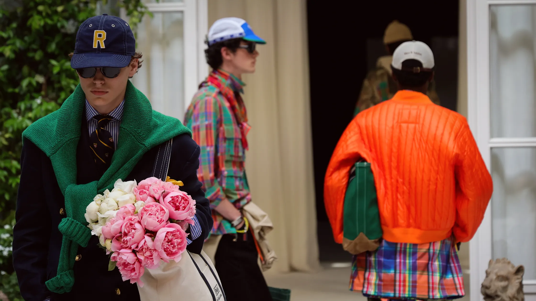 Models in Ralph Lauren Spring 2027 menswear at the Milan show — a navy blazer with green shawl knit and a peony bouquet, layered madras plaid, and an orange cable-knit bomber over a plaid kilt