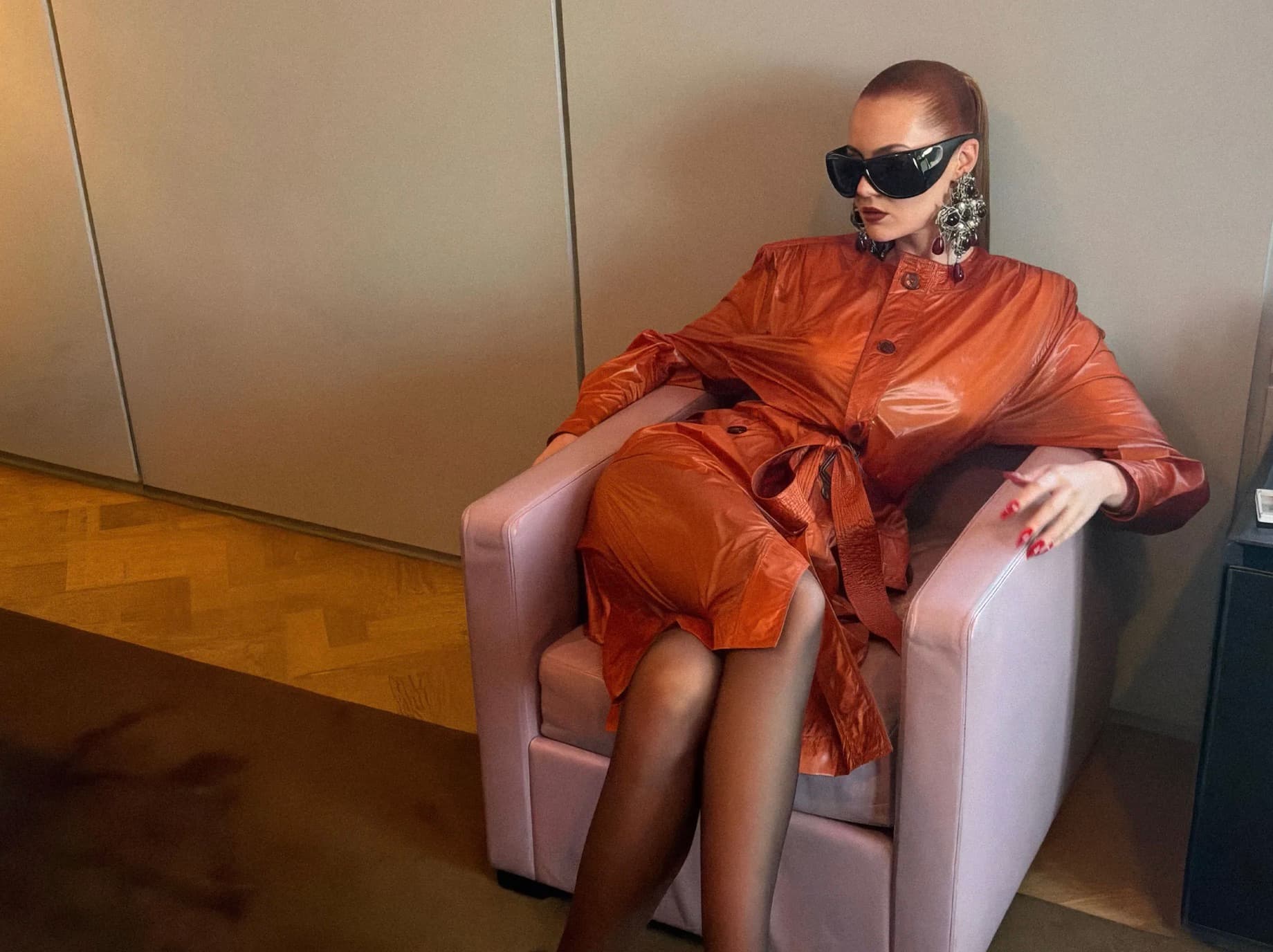 Model in a glossy orange trench coat and sunglasses seated in a lounge chair