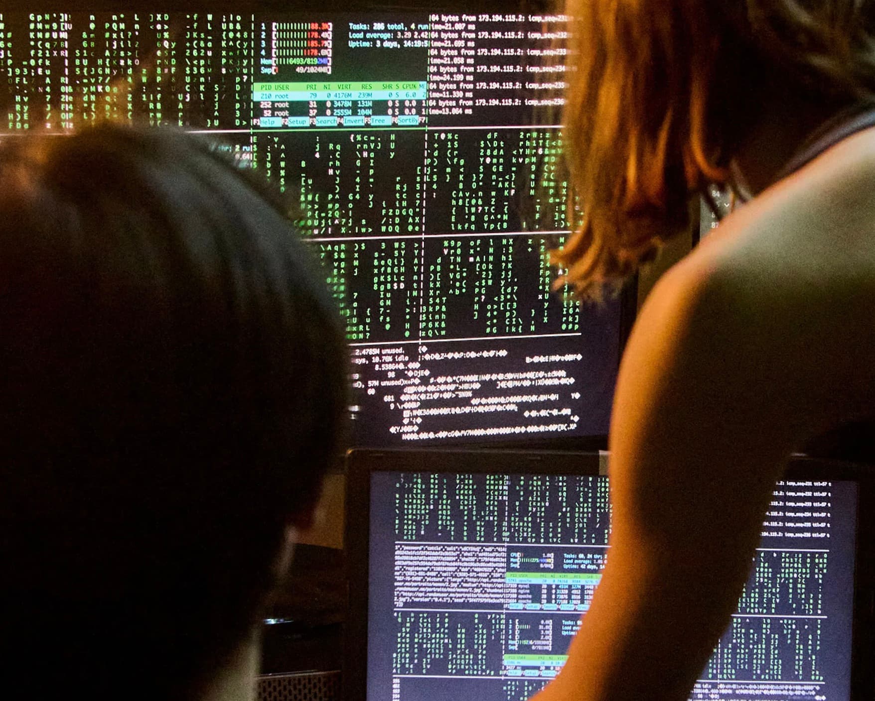 Two people reviewing server monitoring dashboards filled with terminal data on multiple screens