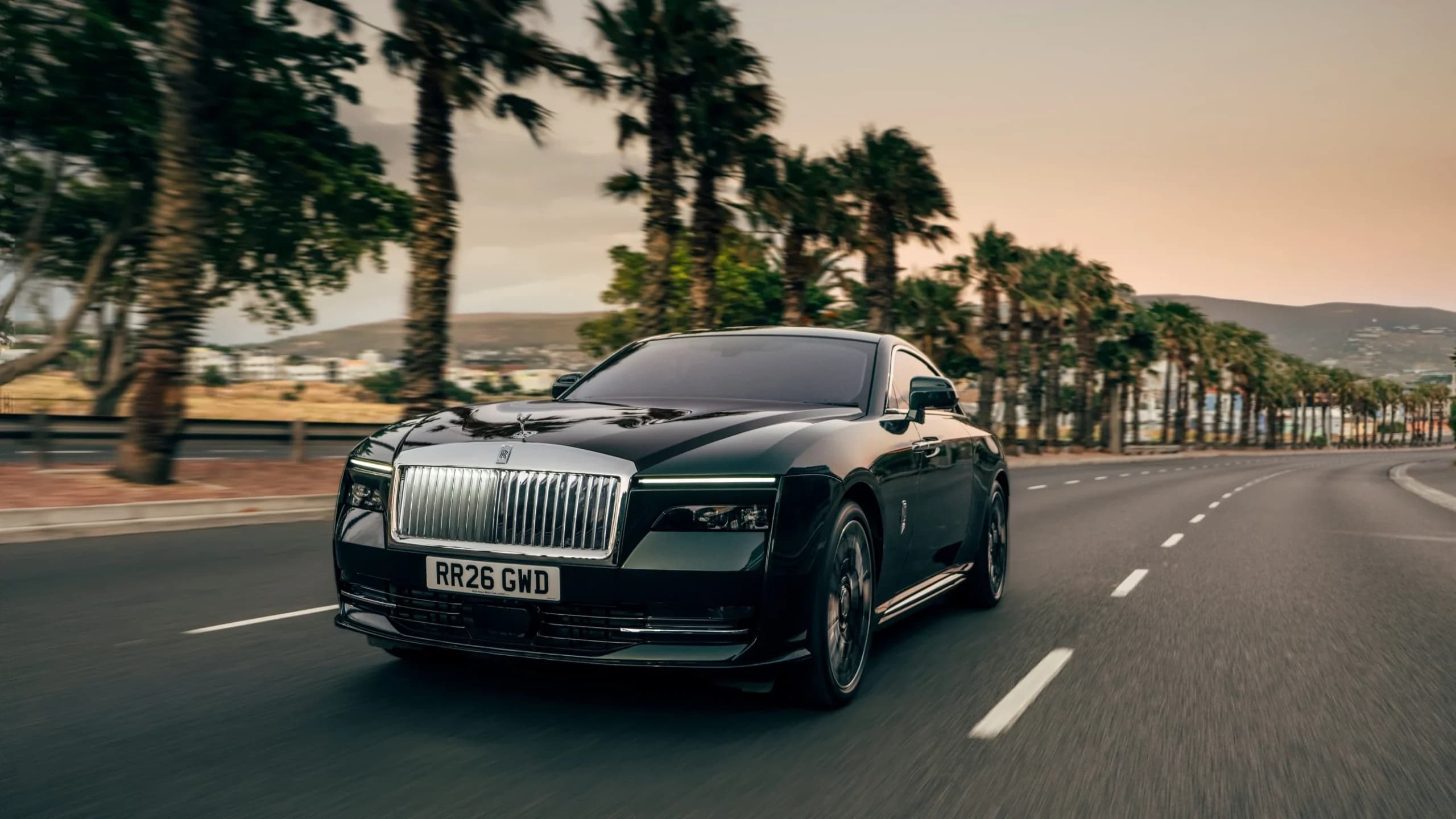 A black Rolls-Royce Spectre Series II electric coupe driving along a palm-lined coastal road at dusk