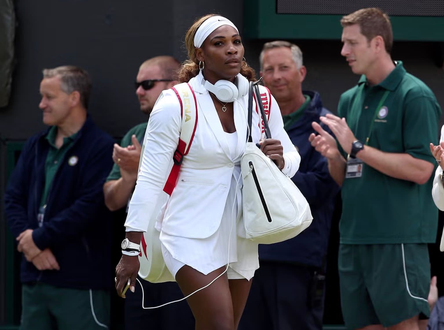 Serena Williams walking onto a Wimbledon court in a white tailored blazer over tennis whites, with headphones around her neck and a Wilson bag on her shoulder