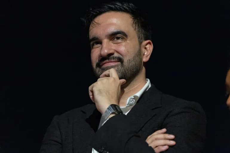 New York City mayor Zohran Mamdani, arms crossed and hand to his chin, smiling at an event