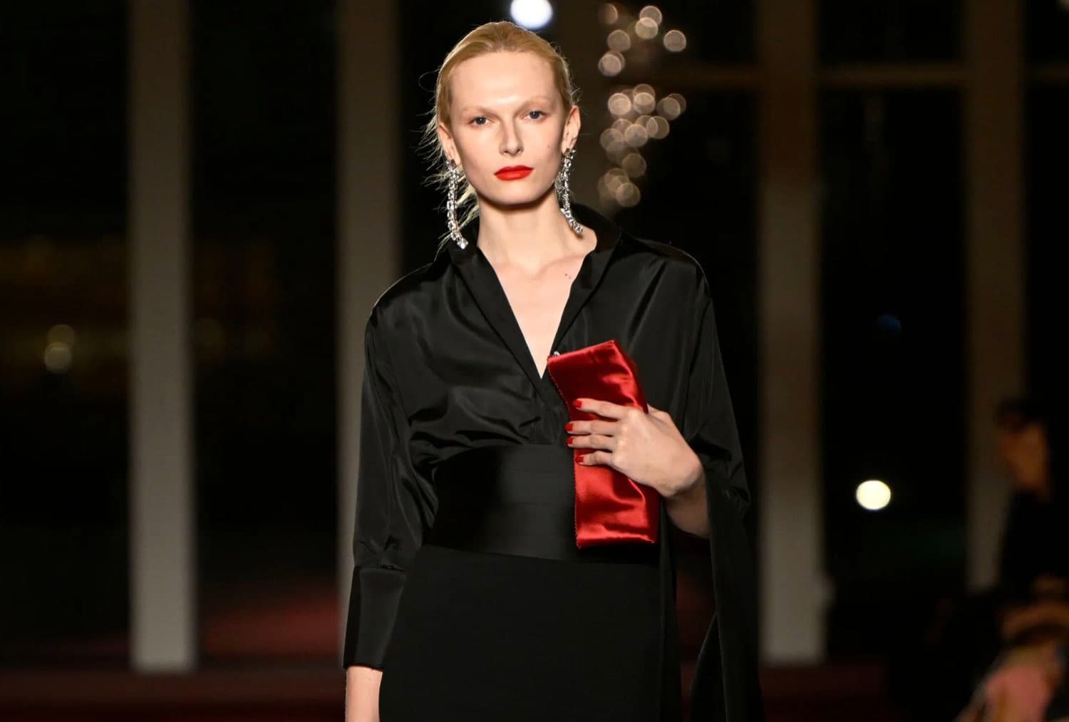 A model on the runway in a black satin gown holding a red clutch