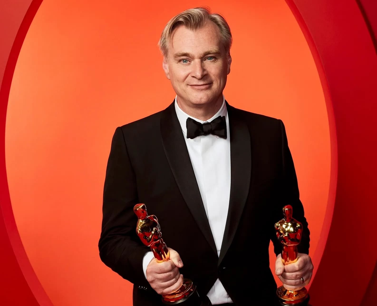 Christopher Nolan in a tuxedo holding two Academy Awards against a red backdrop