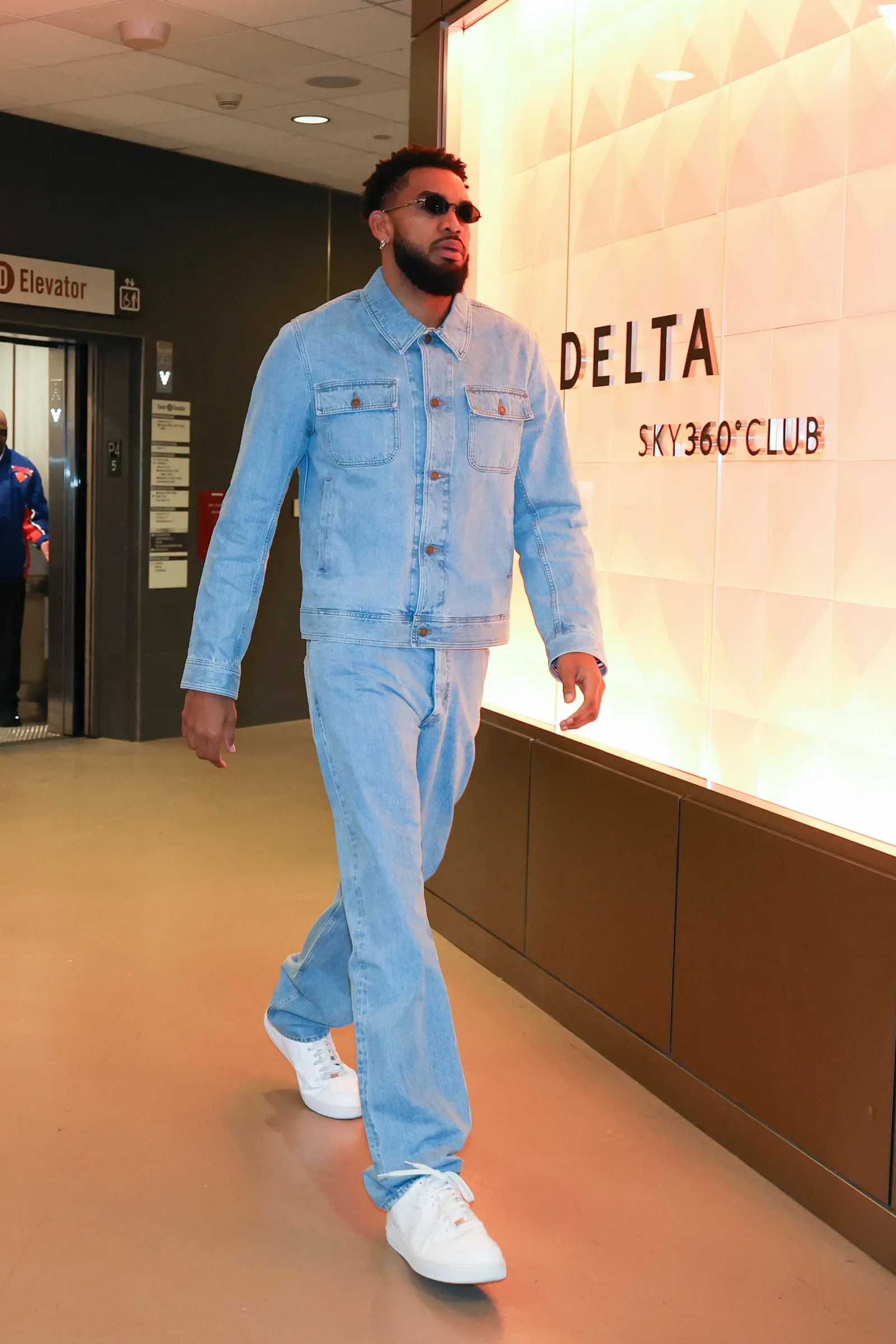 Karl-Anthony Towns walking through an arena hallway in a light denim jacket and matching jeans