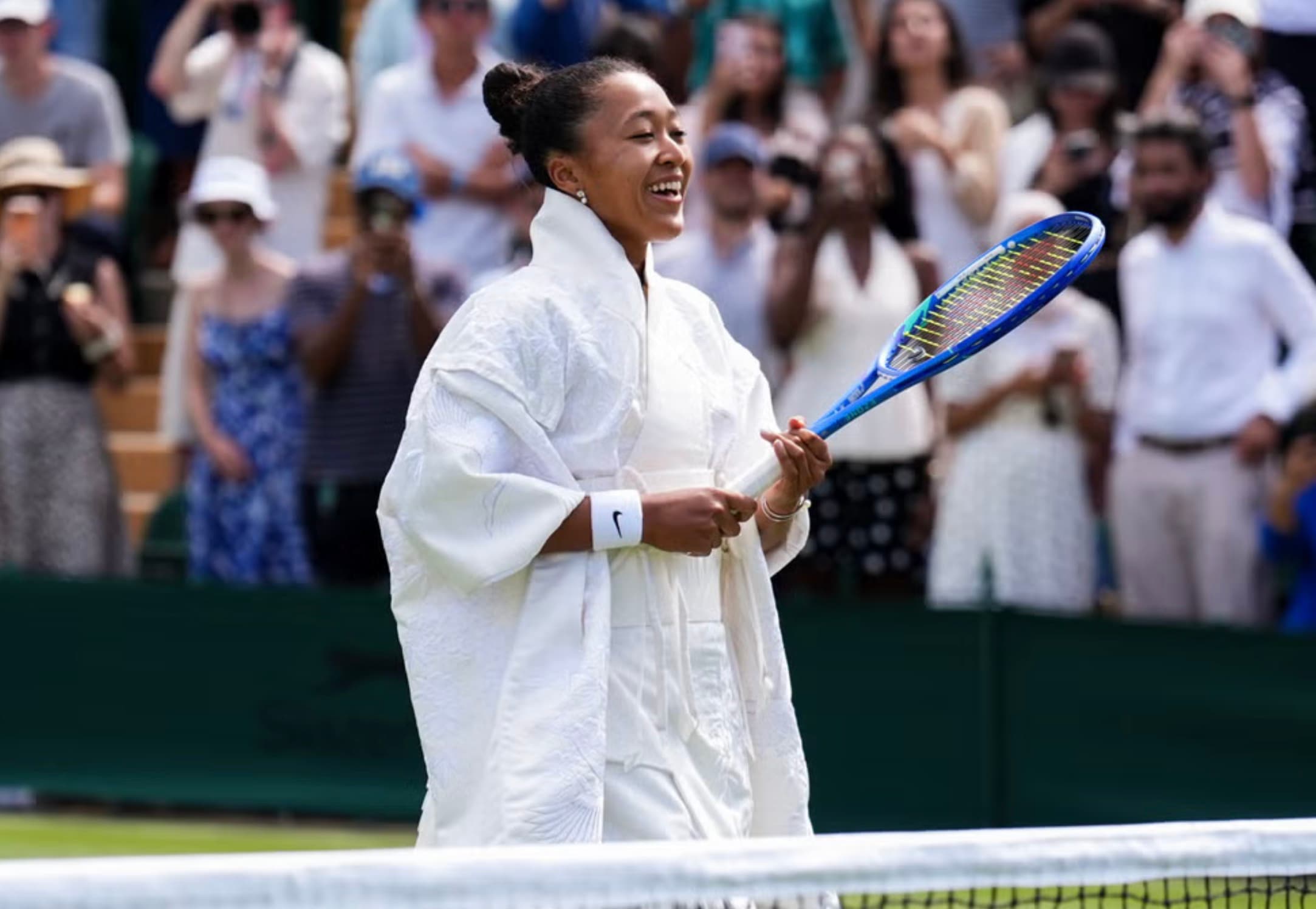 Naomi Osaka smiling on a Wimbledon court in a white kimono inspired robe, holding her racket