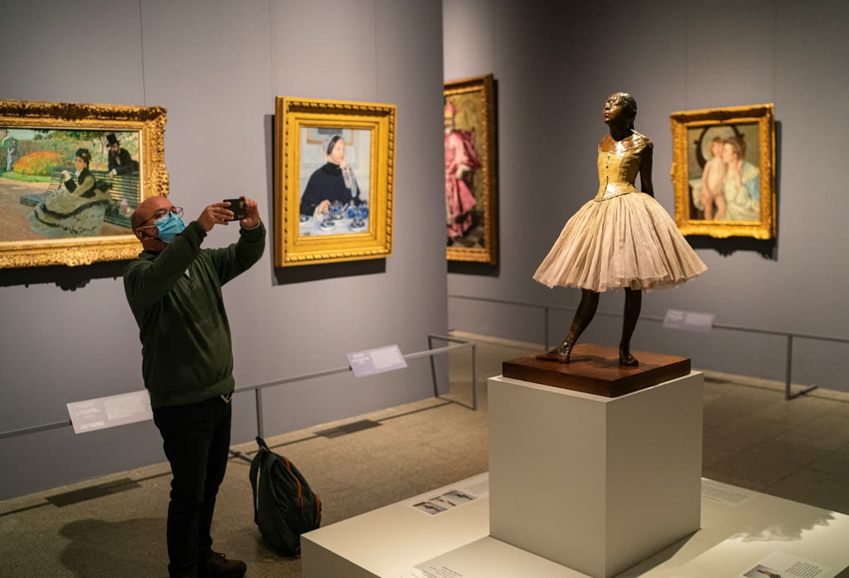 A visitor photographs Degas's Little Dancer sculpture in a museum gallery hung with Impressionist paintings