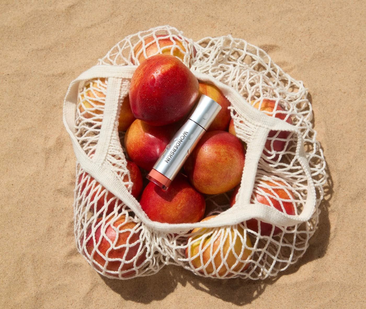 A Wonderskin lip product resting on nectarines in a string bag on sand